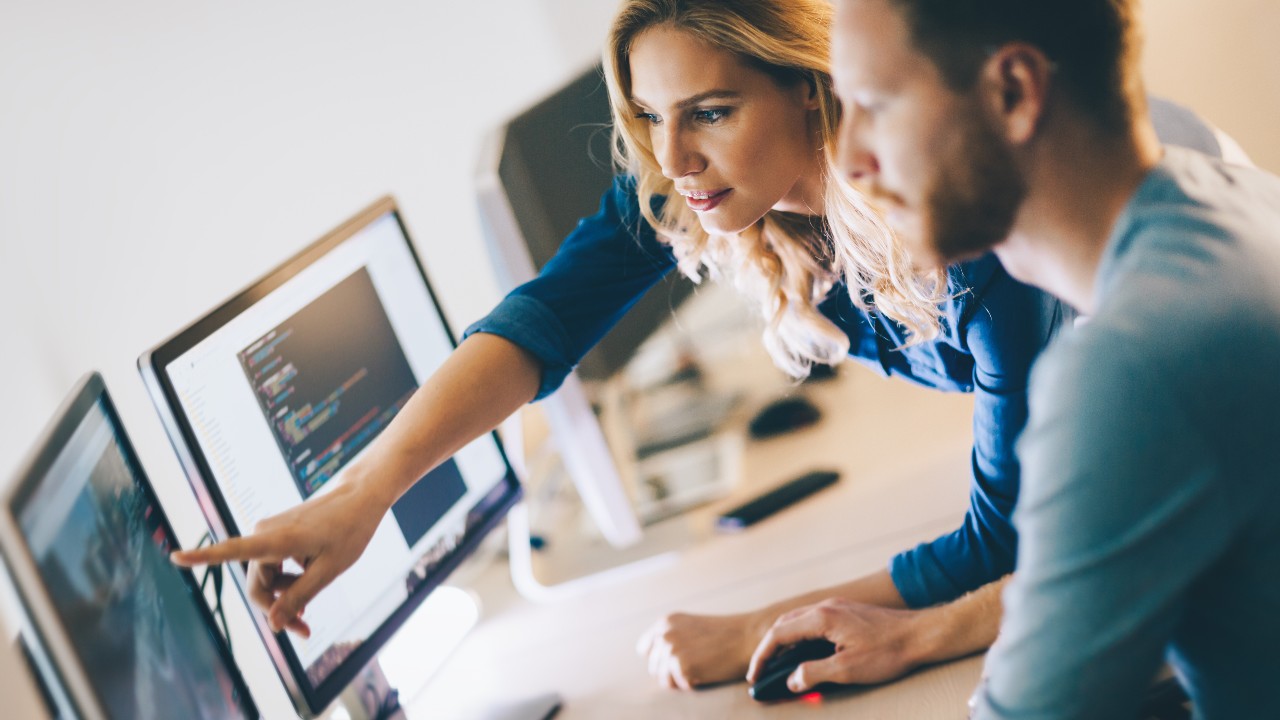 Eine Frau zeigt mit ihrem Finger auf einen PC Monitor und ein Mann Vordergrund schaut in die gezeigte Richtung 